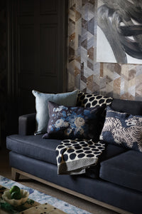 A moodily shot corner of a room with a lustrous black sofa.  On the sofa sits an array of plump blue cushions.