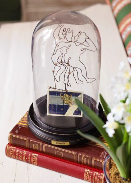 A clear glass dome display case with a black base, featuring a line-art zodiac constellation design and a small Gemini-themed card, styled on stacked vintage books with white flowers in the foreground.