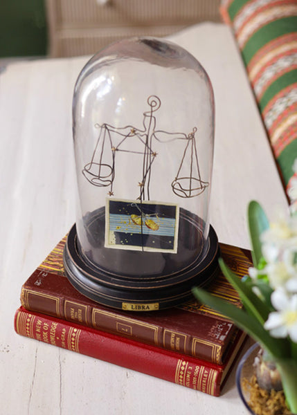A clear glass dome cloche with a black base displaying a Libra zodiac symbol made from wire and a small card, resting on two vintage red books on a white tabletop.