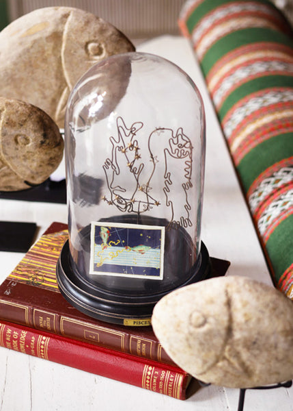 A clear glass dome display case with a black base, decorated with a wire depiction of Pisces, along with an illustrated playing card, showing the astrological constellation, displayed on top of vintage red books with stone fish decor around it.