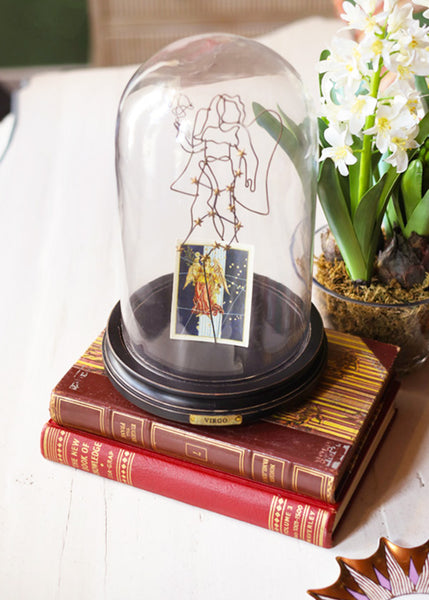 A clear glass dome display with a black circular base sits on top of two vintage red books. Inside the dome is a line-art constellation drawing and a small illustrated card, both depicting the constellation and star sign Virgo.