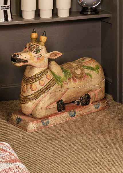 A large hand-painted wooden cow statue with traditional Indian motifs, resting on a textured beige carpet in front of a dark grey shelf.