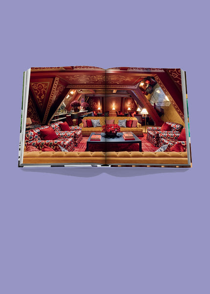 An open hardcover book, Ecclectic, by Sig Bergamin, published by Assouline, displayed against a solid lavender background, showing a wide interior photograph of an ornate lounge with red patterned sofas, warm lighting, and rich painted wood panelling.