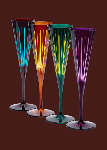 Four elegant L'Objet champagne flutes with a flared shape and vertical ridges, shown in 4 different colours: amber, burgundy and blue, green and purple against a solid dark red background. 