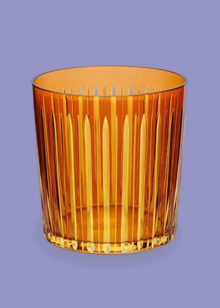An amber-colored L'Objet Prism glass tumbler with a vertical fluted pattern, shown against a solid lavender background.