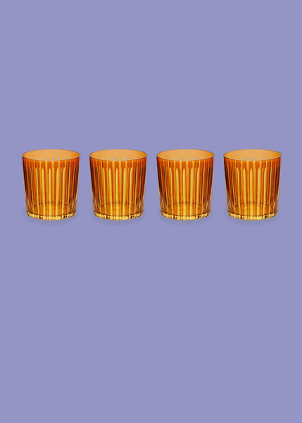 Four amber-colored L'Objet ribbed glass tumblers arranged in a row against a solid lavender background.