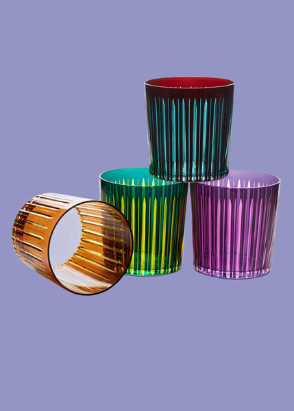 Four L'Objet ribbed glass tumblers in amber, green, purple, and teal red, arranged on a solid lavender background.