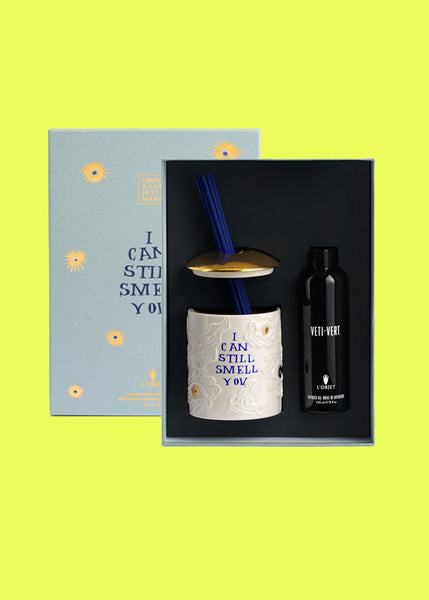 A gift set featuring a Ruan Hoffman x L'Objet ceramic diffuser with blue reeds, a black bottle of Veti-vert diffuser liquid, and a light blue decorative box with yellow floral accents, all arranged on a bright yellow background.
