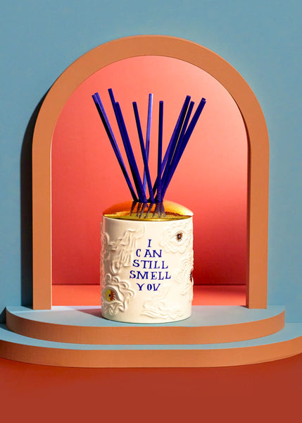 A L'Objet Ruan Hoffmann ceramic reed diffuser with embossed detailing and the text 'I CAN STILL SMELL YOU' in blue, filled with blue reeds, displayed on a tiered platform inside a peach arch against a blue and coral background.