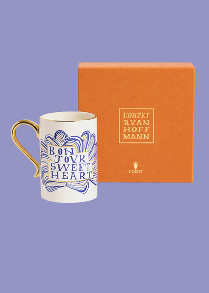 A square orange gift box with the words "L'Objet Ruan Hoffmann" in the centre sits behind a white ceramic mug.  The mug has a gold handle and the words "bonjour sweetheart"