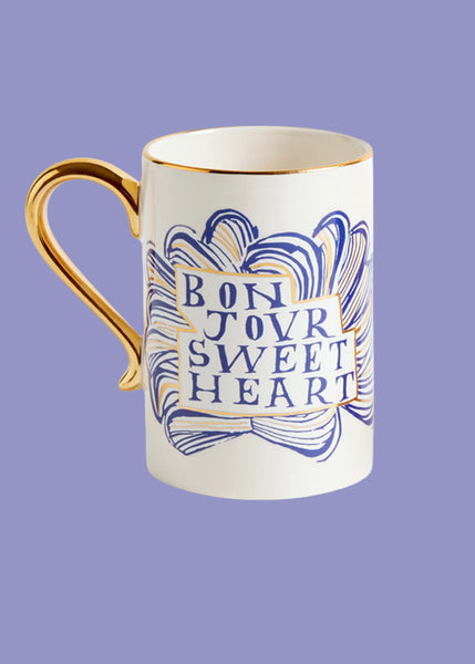 A white ceramic mug with a gold handle bears the words "bonjour sweetheart"  Hand painted abstract blue ribbon illustrations form a cloud chape behind the text.