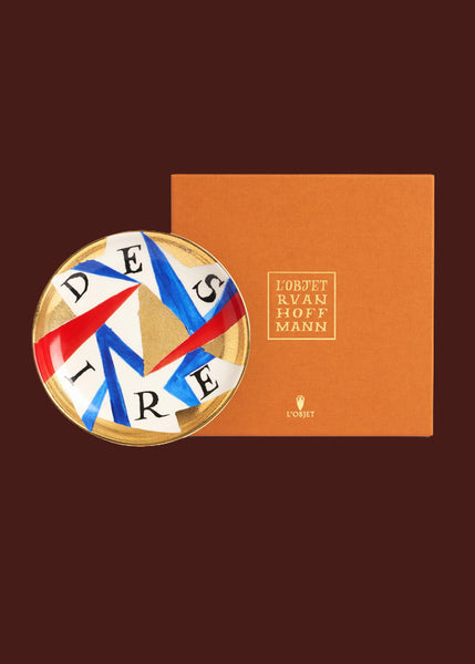 A colourful plate with gold details sits against an orange luxury gift box bearing the words "Lo'Objet Ruan Hoffmann"  The word "desire" is hand painted on this red, gold, blue and white hand made round ceramic plate. 