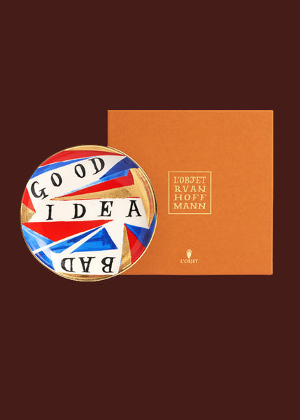 A colourful plate with gold details sits against an orange luxury gift box bearing the words "Lo'Objet Ruan Hoffmann"  The words "Good idea bad" are hand painted on this red, gold, blue and white hand made round ceramic plate. 