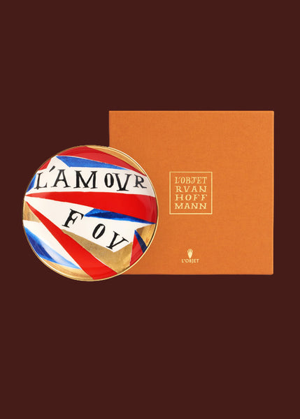 A colourful plate with gold details sits against an orange luxury gift box bearing the words "Lo'Objet Ruan Hoffmann"  The words "L'amour Fou" are hand painted on this red, gold, blue and white hand made round ceramic plate. 
