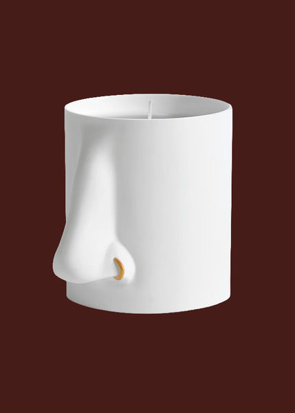 A white sculptural candle votive shaped like a stylised nose, shown against a solid dark red background.